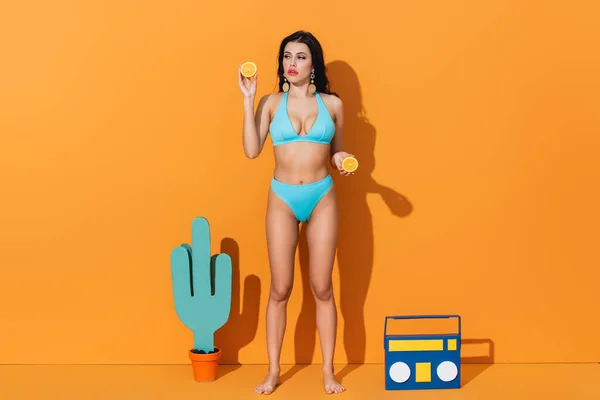 Frau im Badeanzug hält orangefarbene Hälften, während sie in der Nähe von Papierkaktus und Boombox auf Orange steht — Stockfoto