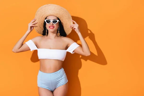 Стильна дівчина в сонцезахисних окулярах посміхається і торкається солом'яного капелюха на помаранчевому — стокове фото