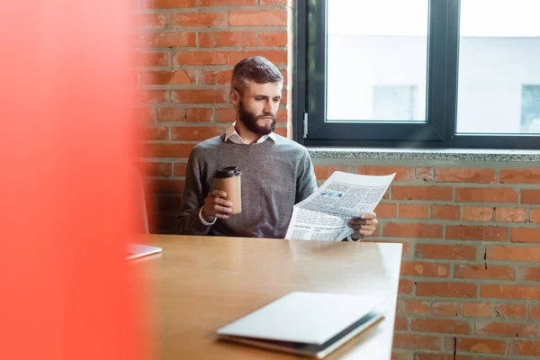 Selektiver Fokus eines bärtigen Geschäftsmannes, der Coffee to go in der Hand hält und neben Laptop Zeitung liest — Stockfoto