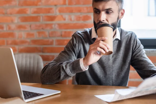 Enfoque selectivo de hombre de negocios guapo mirando a la computadora portátil mientras sostiene periódico y beber café para ir - foto de stock
