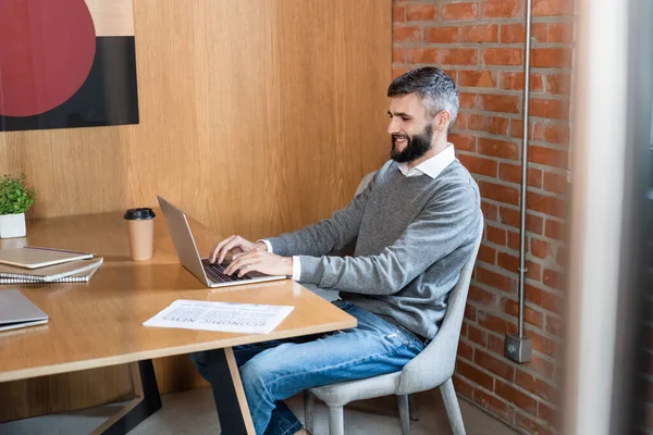 Selektiver Fokus eines gut aussehenden Geschäftsmannes, der lächelnd Laptop in der Nähe von Zeitung und Pappbecher benutzt — Stockfoto