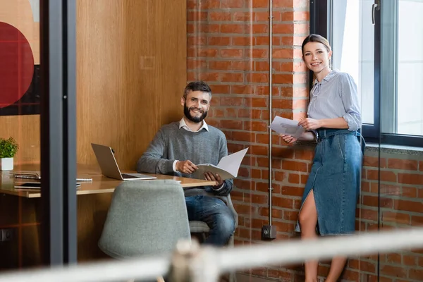 Selective focus of happy businesswoman and businessman holding documents and looking at camera in office — Stock Photo