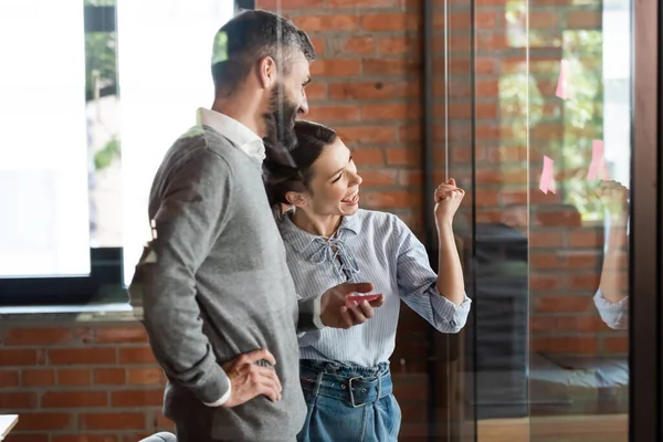 Selektiver Fokus aufgeregter Geschäftsfrau gestikuliert beim Betrachten klebriger Zettel auf Glas in der Nähe von Geschäftsleuten — Stockfoto