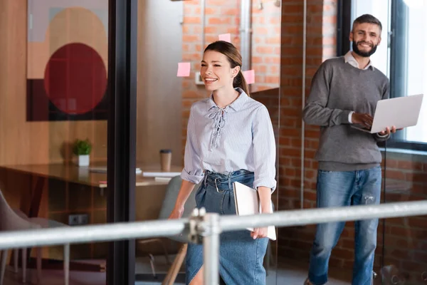 Selektiver Fokus einer fröhlichen Geschäftsfrau, die mit Laptop in der Nähe eines bärtigen Geschäftsmannes spaziert — Stockfoto