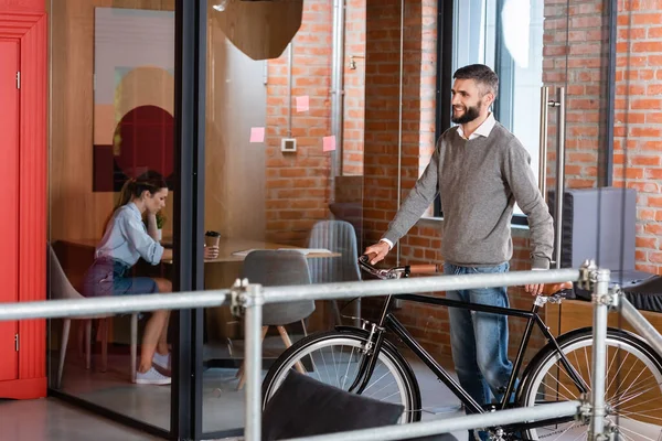 Selective focus of cheerful businessman standing with bicycle near businesswoman sitting in office — Stock Photo