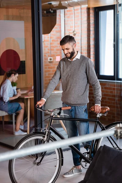 Избирательный фокус счастливого бизнесмена, стоящего на велосипеде рядом с бизнес-леди, сидящей в офисе — стоковое фото