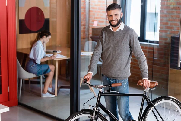 Foco seletivo de empresário alegre de pé com bicicleta perto da mulher sentada no escritório — Fotografia de Stock