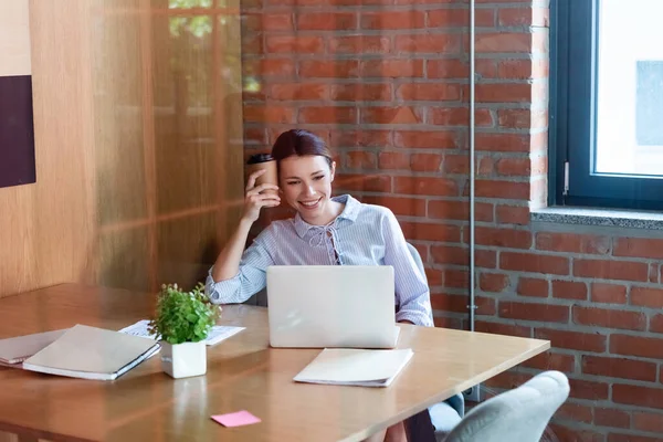 Mulher de negócios feliz olhando para laptop e segurando copo de papel — Fotografia de Stock