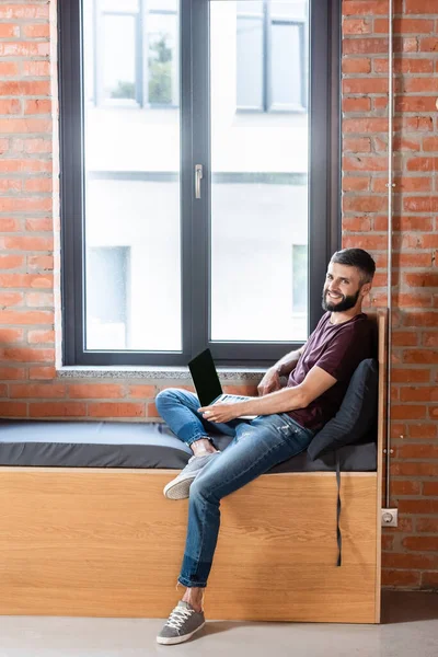 Hombre de negocios feliz usando el ordenador portátil con la pantalla en blanco mientras está sentado en el banco de la ventana en la oficina moderna - foto de stock
