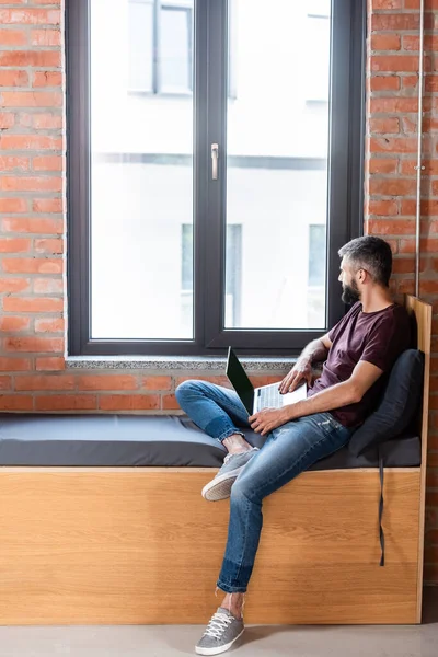 Homem de negócios barbudo usando laptop com tela em branco enquanto sentado no banco de janela no escritório moderno — Fotografia de Stock