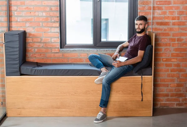 Бородатий бізнесмен сидить з ноутбуком на віконній лавці в сучасному офісі — стокове фото