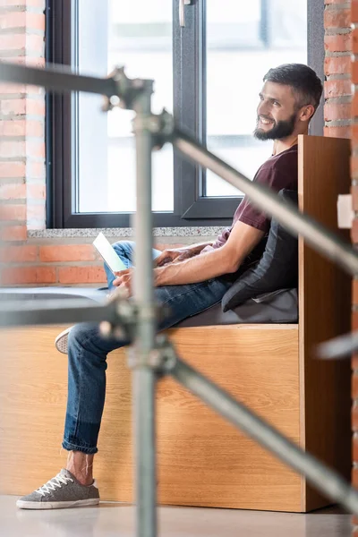Избирательный фокус счастливого бизнесмена, сидящего на оконной скамейке и использующего ноутбук — стоковое фото