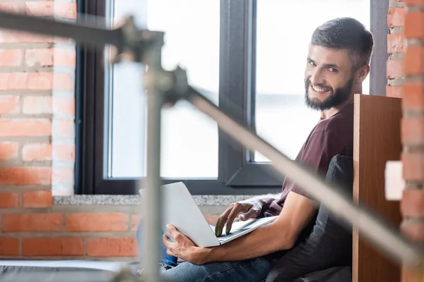 Foco seletivo de empresário alegre sentado no banco de janela e usando laptop — Fotografia de Stock