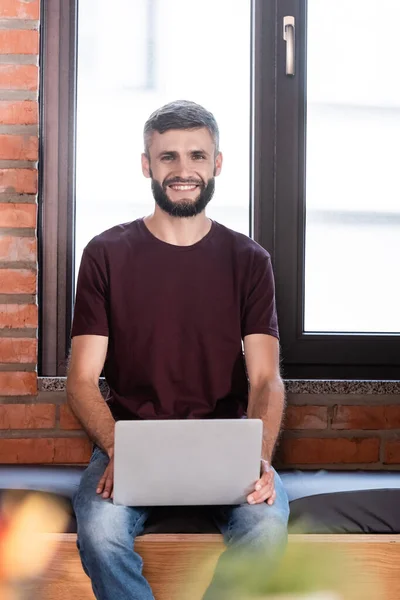 Selektiver Fokus des fröhlichen Geschäftsmannes, der auf der Fensterbank sitzt und Laptop in der Hand hält — Stockfoto