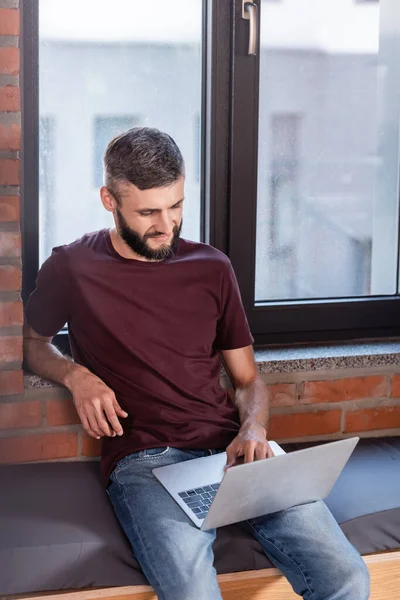 Bearded businessman sitting on window bench and using modern laptop — Stock Photo