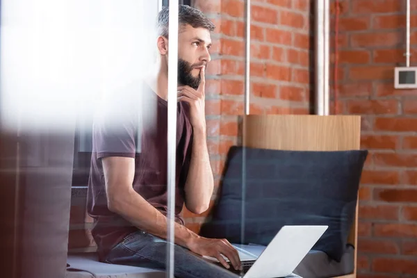 Enfoque selectivo de empresario pensativo sentado en el banco de la ventana y el uso de un ordenador portátil moderno - foto de stock