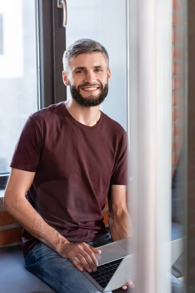 Selective focus of cheerful businessman sitting on window bench and holding modern laptop while looking at camera — Stock Photo