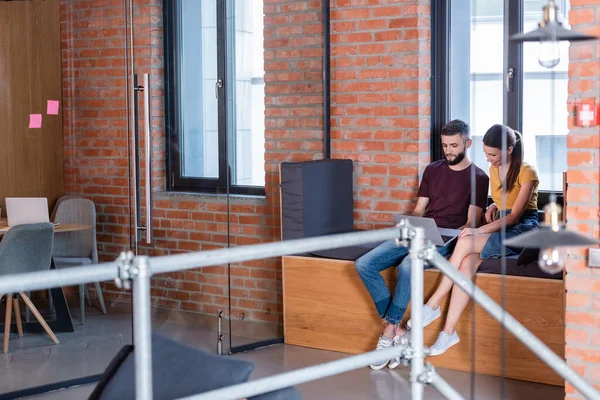 Selective focus of businesswoman and bearded businessman using laptop while sitting on window bench in office — Stock Photo