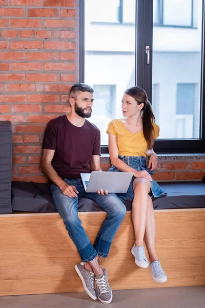 Happy businesswoman and bearded businessman using laptop while sitting on window bench in office — Stock Photo