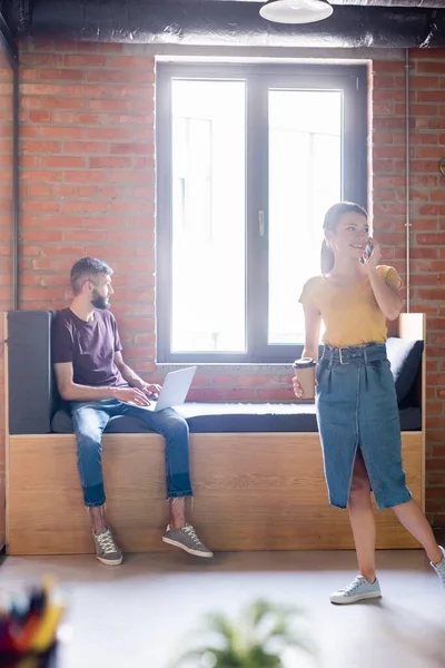 Enfoque selectivo de la mujer de negocios feliz hablando en el teléfono inteligente y sosteniendo taza de papel cerca de hombre de negocios utilizando el ordenador portátil - foto de stock