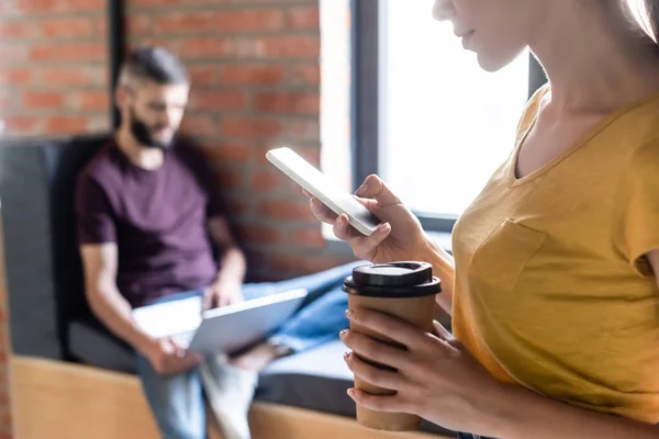 Selective focus of businesswoman using smartphone and holding paper cup near businessman with laptop in office — Stock Photo