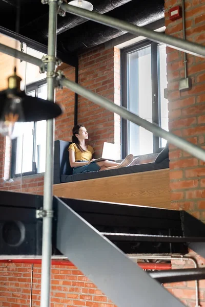Selektiver Fokus der Geschäftsfrau, die mit Laptop auf der Fensterbank sitzt und im Büro Fenster betrachtet — Stockfoto
