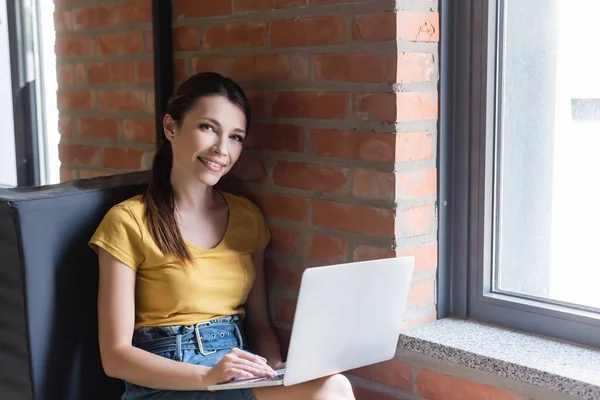 Mujer de negocios sonriente sentada en el banco de la ventana y usando el ordenador portátil en la oficina - foto de stock