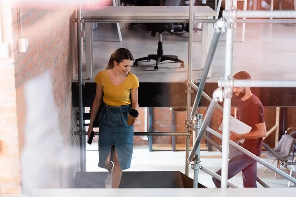 Enfoque selectivo de la mujer de negocios feliz sosteniendo taza de papel y teléfono inteligente cerca de hombre de negocios mientras camina por las escaleras - foto de stock