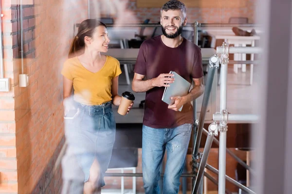 Избирательный фокус веселой деловой женщины, держащей бумажный стаканчик и смартфон рядом с бизнесменом с ноутбуком во время прогулки по лестнице — стоковое фото