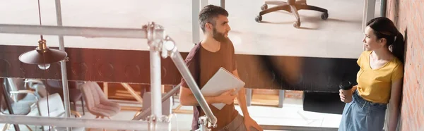 Панорамный снимок предпринимательницы, держащей бумажный стаканчик и смотрящей на бизнесмена с ноутбуком — стоковое фото