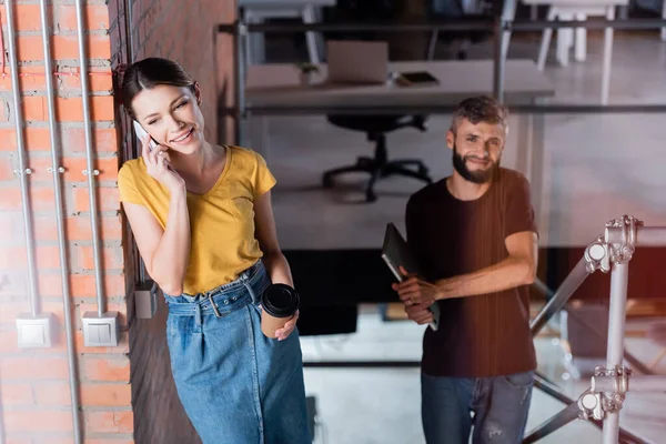 Selektiver Fokus glücklicher Geschäftsfrau, die in der Nähe eines bärtigen Geschäftsmannes mit Laptop auf dem Smartphone spricht — Stockfoto