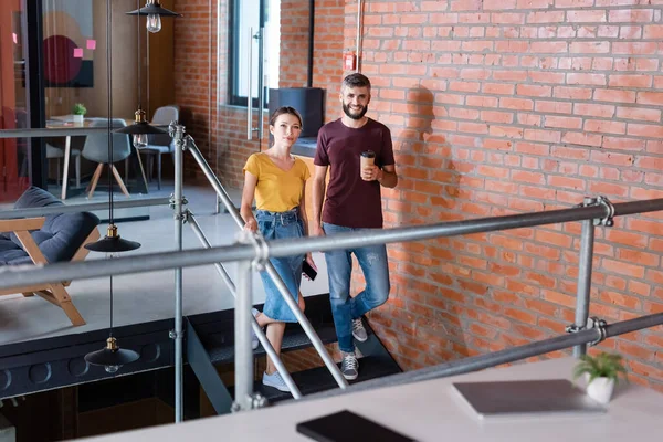 Enfoque selectivo de atractiva empresaria sosteniendo teléfono inteligente cerca de hombre de negocios con taza de papel caminando en las escaleras - foto de stock