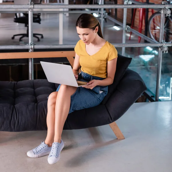 Atractiva mujer de negocios usando el ordenador portátil mientras está sentado en el sofá en la oficina moderna - foto de stock