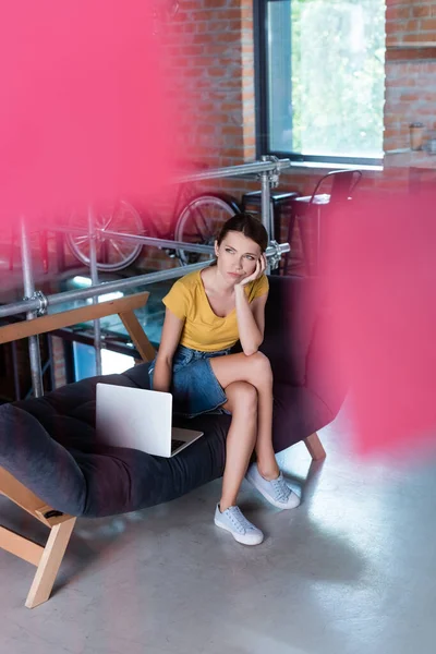 Enfoque selectivo de la mujer de negocios pensativa mirando hacia otro lado mientras está sentado cerca de la computadora portátil en el sofá - foto de stock