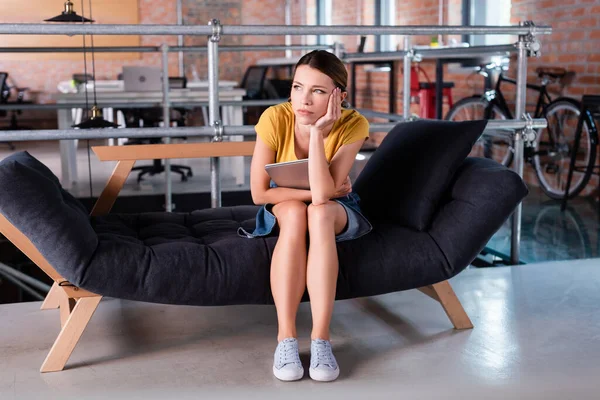 Pensativa mujer de negocios mirando hacia otro lado mientras está sentado en el sofá y la celebración de ordenador portátil - foto de stock