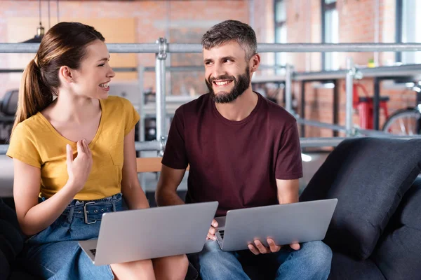 Happy businessman and cheerful businesswoman looking at each other near laptops while sitting on sofa — Stock Photo