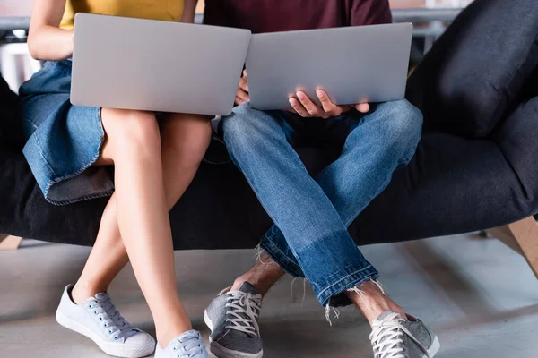 Vista recortada de hombre de negocios y mujer de negocios usando computadoras portátiles mientras está sentado en el sofá - foto de stock