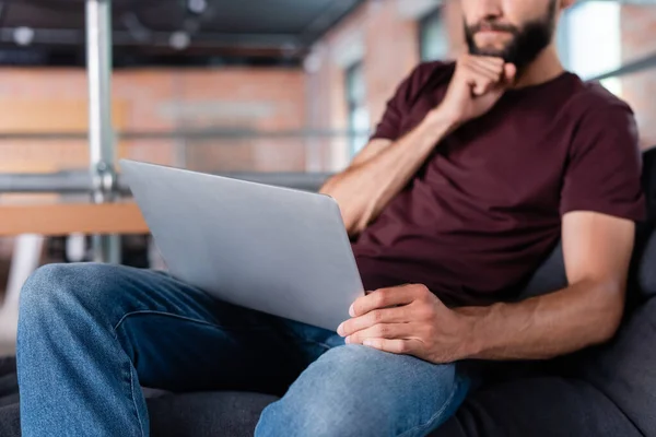 Vista ritagliata di pensieroso uomo d'affari pensare e toccare il computer portatile mentre seduto sul divano in ufficio — Foto stock