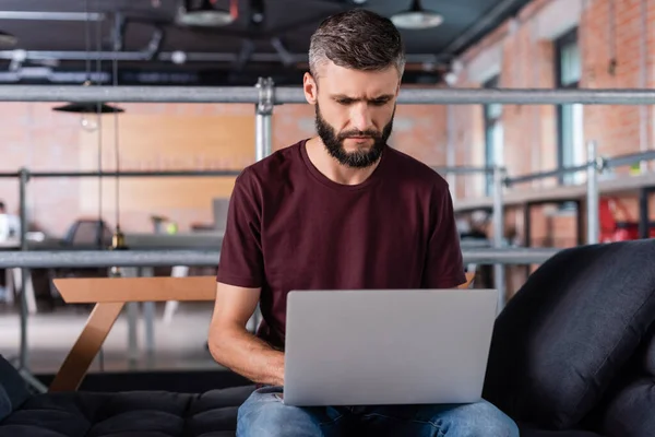 Homme d'affaires concentré assis sur le canapé et utilisant un ordinateur portable dans le bureau — Photo de stock