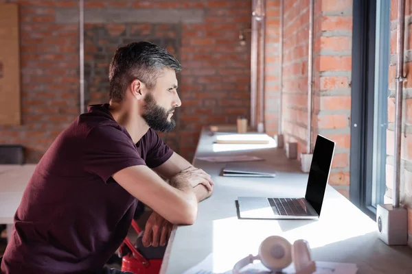 Side view of bearded businessman looking at laptop near wireless headphones — Stock Photo