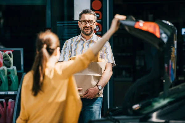Foco seletivo de homem sorridente segurando saco de compras e olhando para a mulher perto do carro na rua urbana — Fotografia de Stock