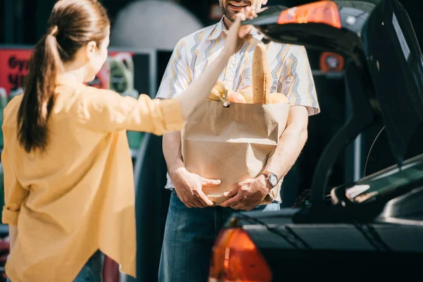 Foco seletivo de homem sorridente segurando saco de compras com comida perto de esposa e carro tronco na rua urbana — Fotografia de Stock