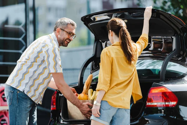 Foco seletivo da mulher abrindo o porta-malas do carro perto de homem sorridente segurando saco de compras com comida na rua urbana — Fotografia de Stock