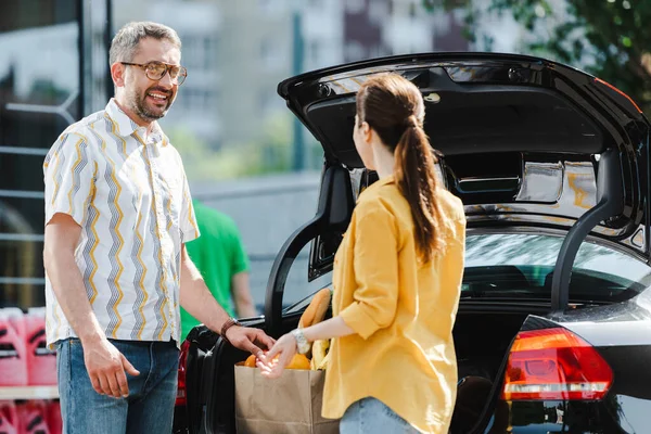 Selective focus of smiling man looking at woman near open car trunk on urban street — Stock Photo