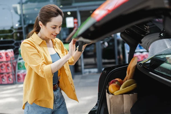 Selektiver Fokus einer lächelnden Frau, die neben einer Einkaufstasche mit Lebensmitteln im Kofferraum eines Autos auf einer städtischen Straße steht — Stockfoto