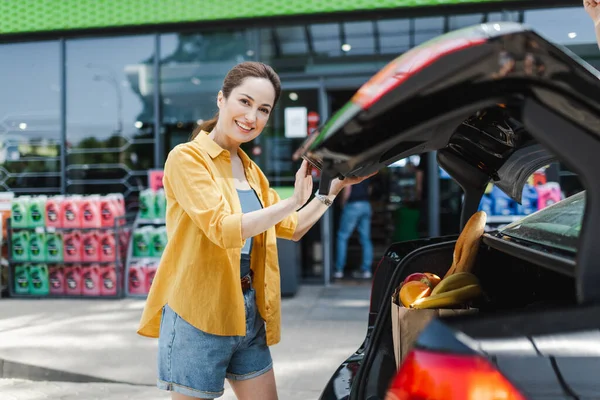 Concentration sélective de la femme souriant à la caméra tout en se tenant près du sac à provisions avec de la nourriture dans le coffre de la voiture dans la rue urbaine — Photo de stock