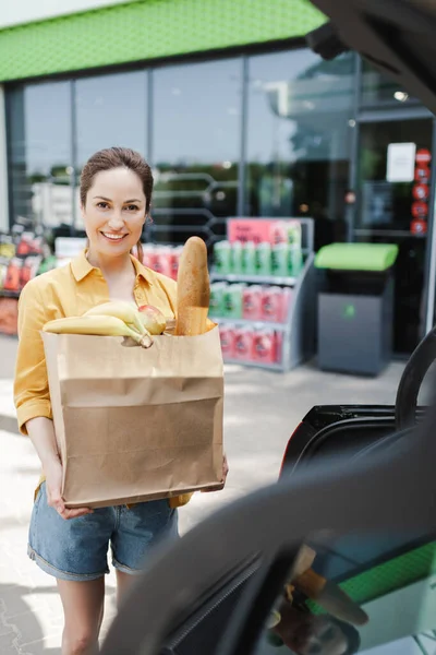 Focus selettivo della donna sorridente che tiene la borsa della spesa con cibo vicino all'auto sulla strada urbana — Stock Photo