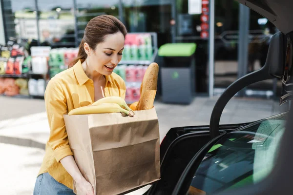 Selective focus of smiling woman holding shopping bag with food near open car trunk on urban street — Stock Photo