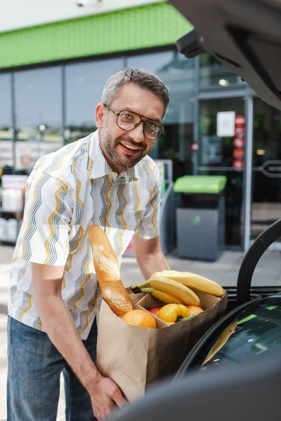 Selektiver Fokus eines lächelnden Mannes, der eine Einkaufstasche mit Lebensmitteln in den offenen Kofferraum eines Autos auf einer städtischen Straße legt — Stockfoto