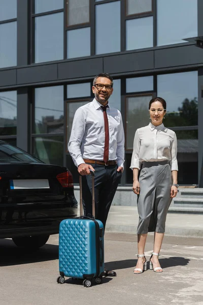 Smiling businessman holding suitcase near businesswoman and car on urban street — Stock Photo